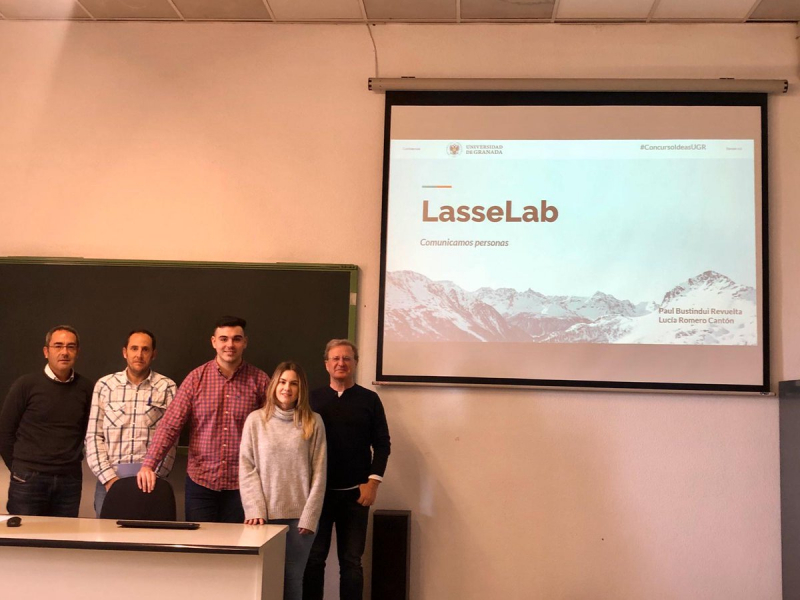 Grupo proyecto LasseLab Paul Bustindui y Lucía Romero junto con 3 personas más en una clase y en la pizarra digital la portada de su proyecto LasseLab